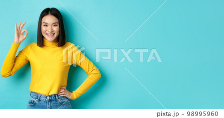 Happy beautiful asian woman smiling, showing okay gesture in approval, saying yes, standing pleased over blue background 98995960
