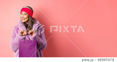 Shopping and fashion concept. Fashionable gradmother bought present and smiling, holding purple bag in hands, standing over pink background Shopping and fashion concept. Fashionable gradmother bought present and smiling, holding purple bag in hands, standing over pink background 98995978
