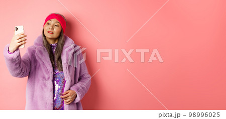 Fashion concept. Stylish asian senior female taking selfie on smartphone, posing in purple faux fur coat and party dress, standing over pink background 98996025