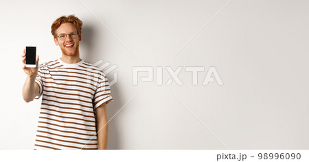 Technology and e-commerce concept. Happy young redhead man in glasses showing blank smartphone screen, smiling satisfied, standing over white background Technology and e-commerce concept. Happy young redhead man in glasses showing blank smartphone screen, smiling satisfied, standing over white background 98996090