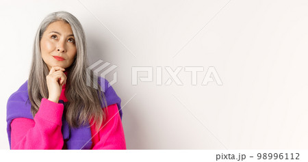 Close up of thoughtful asian female with grey hair, looking up and thinking, making decision, standing over white background Close up of thoughtful asian female with grey hair, looking up and thinking, making decision, standing over white background 98996112