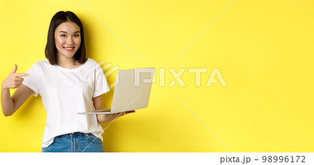 Smiling asian woman pointing finger at her laptop, showing something online, standing over yellow background 98996172
