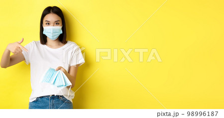 Coronavirus, quarantine and medicine concept. Young asian woman pointing finger at medical masks for going outdoors, standing over yellow background 98996187