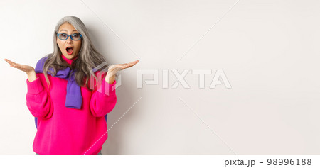 Fashionable asian senior woman in sunglasses gasping amazed, spread hands sideways and staring surprised at camera, white background 98996188