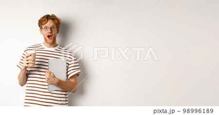 Amazed redhead man talking with coworkers on coffee break, holding cup and laptop, staring surprised at camera, white background 98996189