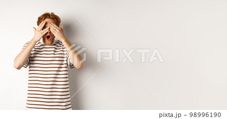 Shocked and embarrassed redhead guy covering eyes, peeking through fingers and gasping, standing over white background 98996190