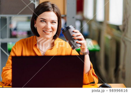 a brunette woman in an orange blouse drinks coffee and works on a laptop a brunette woman in an orange blouse drinks coffee and works on a laptop 98996616