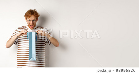 Surprised boyfriend open shopping bag with valentines day gift, staring inside with amazed face, standing over white background 98996826