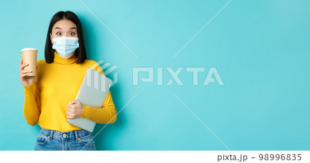 covid-19, health care and quarantine concept. Asian girl student in medical mask standing with laptop and coffee from cafe, standing over blue background covid-19, health care and quarantine concept. Asian girl student in medical mask standing with laptop and coffee from cafe, standing over blue background 98996835