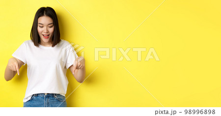 Beauty and fashion concept. Beautiful asian woman in white t-shirt pointing fingers down, demonstrate logo standing over yellow background Beauty and fashion concept. Beautiful asian woman in white t-shirt pointing fingers down, demonstrate logo standing over yellow background 98996898