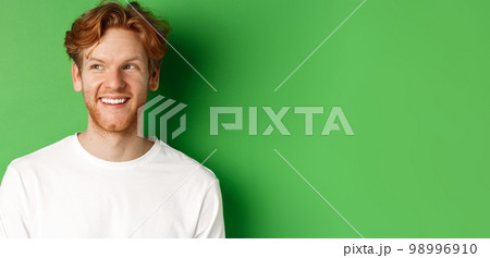 Headshot of happy redhead man with beard wearing white long sleeve, looking left at copy space and smiling, standing over green background 98996910