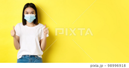 Covid-19, quarantine and social distancing concept. Cheerful asian woman in medical mask and white t-shirt showing thumbs up in approval, praise good deal, yellow background 98996918