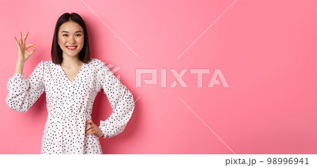 Pretty young asian woman in dress showing okay sign, praising and showing approval, looking satisfied, standing against pink background Pretty young asian woman in dress showing okay sign, praising and showing approval, looking satisfied, standing against pink background 98996941