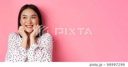 Beauty and skin care concept. Close-up of beautiful asian woman with clean face, blushing and looking dreamy left, admiring promo offer, standing over pink background 98997299