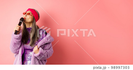 Stylish asian senior woman singing song, performing karaoke with microphone, standing in party outfit and faux fur coat against pink background Stylish asian senior woman singing song, performing karaoke with microphone, standing in party outfit and faux fur coat against pink background 98997303