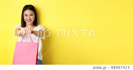 Cute asian girl giving you gift, stretch out hand with pink shopping bag and smiling, congratulating with holiday, standing over yellow background 98997423