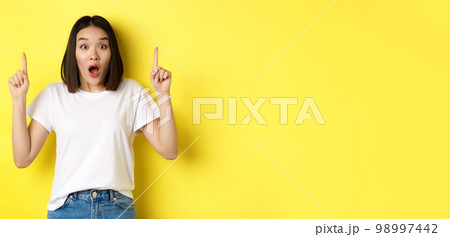 Beauty and fashion concept. Beautiful asian woman in white t-shirt pointing fingers up, standing over yellow background 98997442