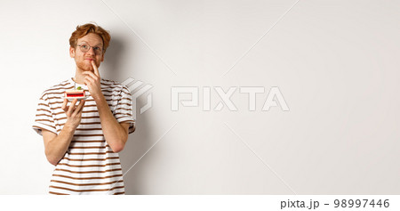 Holidays and celebration concept. Happy young man with red hair and glasses having birthday, holding cake with candle and thinking of b-day wish, standing over white background 98997446
