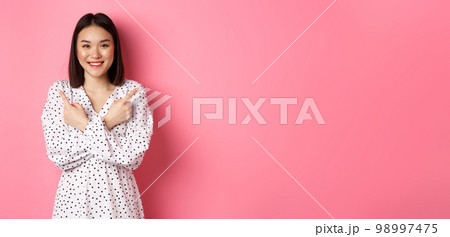 Beautiful asian girl in romantic dress pointing fingers sideways, showing two variants on shopping, smiling at camera, standing against pink background 98997475