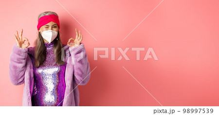 Covid-19, pandemic and fashion concept. Awesome asian grandmother wearing stylish disco dress and respirator, showing okay signs and winking, no proble gesture 98997539