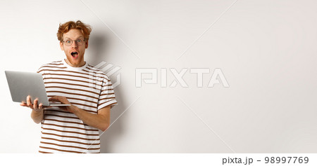 Impressed young man holding laptop in hands, staring at camera with excited face, reading online promotion on website, standing against white backround Impressed young man holding laptop in hands, staring at camera with excited face, reading online promotion on website, standing against white backround 98997769