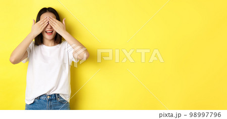 Happy asian woman waiting for surprise gift, close eyes and smiling, expecting something, standing over yellow background Happy asian woman waiting for surprise gift, close eyes and smiling, expecting something, standing over yellow background 98997796