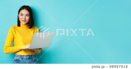 Image of stylish korean woman working on laptop and looking away with dreamy smile, standing with computer against blue background Image of stylish korean woman working on laptop and looking away with dreamy smile, standing with computer against blue background 98997918