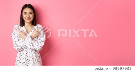 Beautiful asian girl in romantic dress pointing fingers sideways, showing two variants on shopping, smiling at camera, standing against pink background Beautiful asian girl in romantic dress pointing fingers sideways, showing two variants on shopping, smiling at camera, standing against pink background 98997932