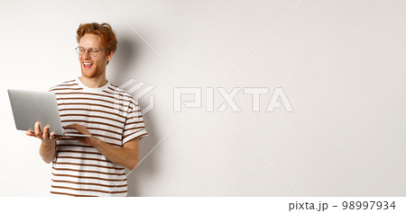 Handsome redhead man working freelance on laptop, smiling and looking pleased at computer screen, white background 98997934