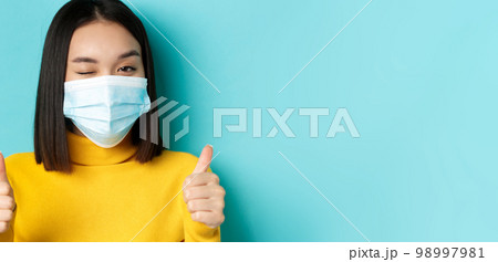 Covid-19, social distancing and pandemic concept. Cute asian girl in medical mask winking at camera, showing thumbs up, good job gesture, praise nice work, blue background 98997981