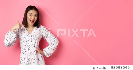 Beautiful asian girl in dress looking happy, pointing finger at herself, standing on romantic pink background 98998049