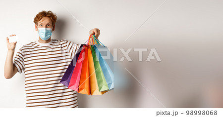Social distancing, coronavirus concept. Young man with red hair, wearing face mask, showing shopping bags and plastic credit card, white background Social distancing, coronavirus concept. Young man with red hair, wearing face mask, showing shopping bags and plastic credit card, white background 98998068