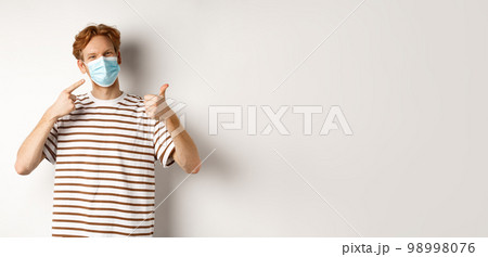 Covid-19, pandemic and social distancing concept. Cheerful young man with red hair pointing at medical mask and showing thumb-up, recommend use preventive measures against virus 98998076