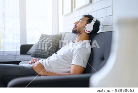 Music, relax and man with headphones on sofa in home living room streaming radio or podcast. Meditation, technology and male on couch in lounge listening to peaceful song, audio or album in house. Music, relax and man with headphones on sofa in home living room streaming radio or podcast. Meditation, technology and male on couch in lounge listening to peaceful song, audio or album in house. 98998663