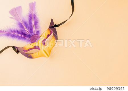 Happy Purim carnival decoration. Top view venetian ball mask with purple feather on pastel background, Jewish Purim and Mardi Gras in Hebrew, holiday background banner design, Masquerade party 98999365