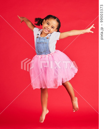Portrait, girl and jumping in tutu skirt on red background, studio and fun fashion. Happy kid, ballet clothes and energy for performance, dance and smile with happiness, ballerina dress and princess 99003373