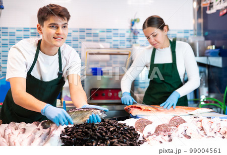 Responsive shop assistant in green apron demonstrating trout in fish store 99004561