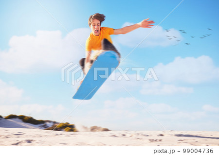 Sport, sandboard man jumping man in a desert for extreme sports, fitness and training against blue sky background. Sandboarding, jump and guy in dunes with energy, freedom and adrenaline in Dubai 99004736