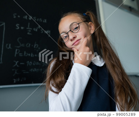Caucasian schoolgirl in glasses answers at the blackboard pointing with a pointer. 99005289