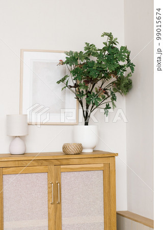 An artificial flower in a pot on a wooden chest of drawers in the living room, decorative items and a frame on the wall in a Scandinavian minimalist style 99015674
