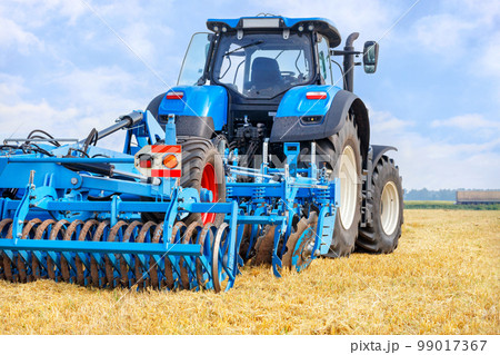 A blue agricultural tractor pulls a harrow across a mowed wheat field on a summer day. 99017367