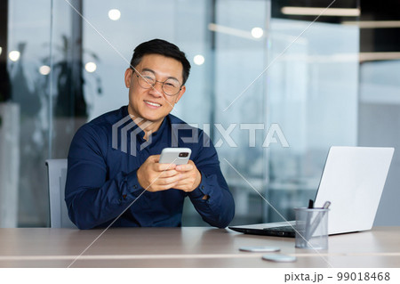 Portrait of successful asian man inside office, businessman in glasses smiling and looking at camera, man at workplace using app on phone and using laptop at work. Portrait of successful asian man inside office, businessman in glasses smiling and looking at camera, man at workplace using app on phone and using laptop at work. 99018468