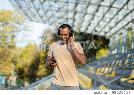 Happy and smiling Indian man in park stadium walking after active jogging and fitness classes, using phone and application for listening to online radio podcasts and music, sportsman in headphones. 99020517
