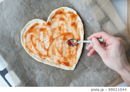 Pizza dough in the shape of a heart, the chef spreads tomato sauce on the pizza. The concept of a surprise for St. Valentine's Day. Pizza dough in the shape of a heart, the chef spreads tomato sauce on the pizza. The concept of a surprise for St. Valentine's Day. 99021044