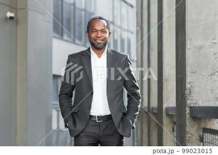 Portrait of a successful African American businessman, director, boss. Standing in a suit outside the office center. He looks confidently into the camera, keeps his hands in his pockets, smiles. 99023015