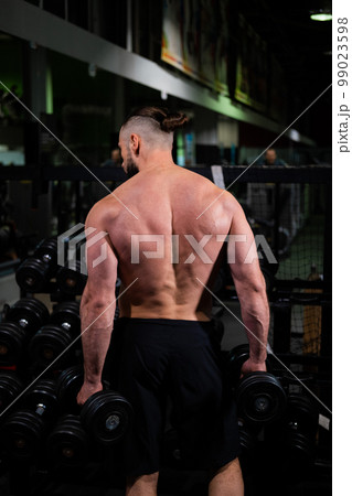 A brutal muscular man with a beard and a perfect body stands with his back and holds dumbbells in his hands in the gym. Fitness, beauty 99023598