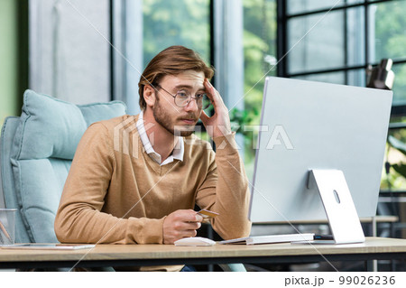Credit card transaction failed. An upset young man sits in the office at the table, looks at the computer, holds a credit card in his hand. Disappointed, he leaned on his arm. 99026236