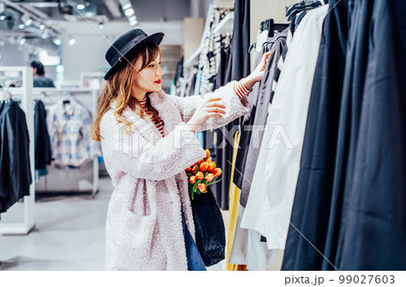 Young fashion woman customer selecting clothes. Buying clothes in boutique using fitting concept while choosing, professional stylist creating trendy look concentrated on selection in designer shop Young fashion woman customer selecting clothes. Buying clothes in boutique using fitting concept while choosing, professional stylist creating trendy look concentrated on selection in designer shop 99027603
