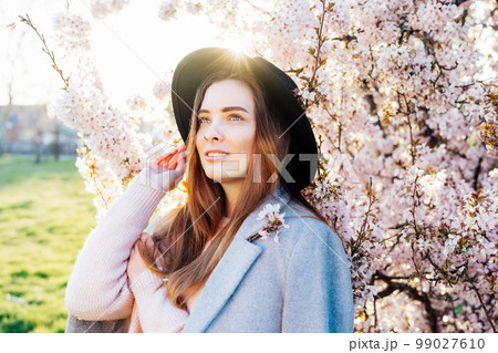 Portrait of a positive, smiling young caucasian woman in hat and coat near a blooming tree with sunset light and sunbeams. Generation Z girl enjoy springtime mood. Selective focus, copy space. Portrait of a positive, smiling young caucasian woman in hat and coat near a blooming tree with sunset light and sunbeams. Generation Z girl enjoy springtime mood. Selective focus, copy space. 99027610
