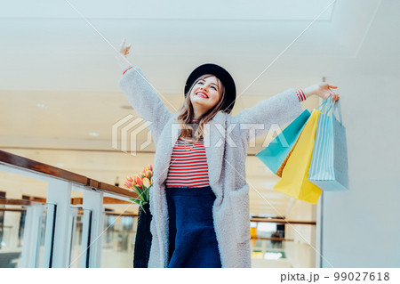 Happy, excited young fashion woman with raised hands holding paper shopping bags on mall background. Woman enjoying new spring season shopping. Consumerism, shopping, sale concept. Selective focus. Happy, excited young fashion woman with raised hands holding paper shopping bags on mall background. Woman enjoying new spring season shopping. Consumerism, shopping, sale concept. Selective focus. 99027618